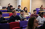 Group of students sit in a lecture theatre on a mixture of coloured chairs.