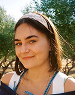 Ellen Allde's face and shoulders. She has brown hair, a headband on and there are trees in the background.