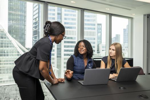 Three women in a meeting room. One is stood up learning over the table speaking to the others. There are office buildings out the window.