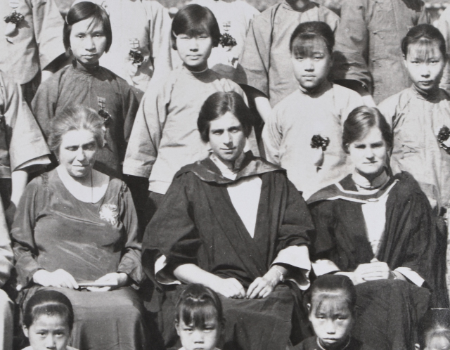 Closeup of a formal group photograph of a school. Three white women are seated centrally, the left two are young women in academic robes and the right an older woman in a dress. Standing behind and sitting in front of the women are Chinese school aged girls in a uniform all wearing corsages.