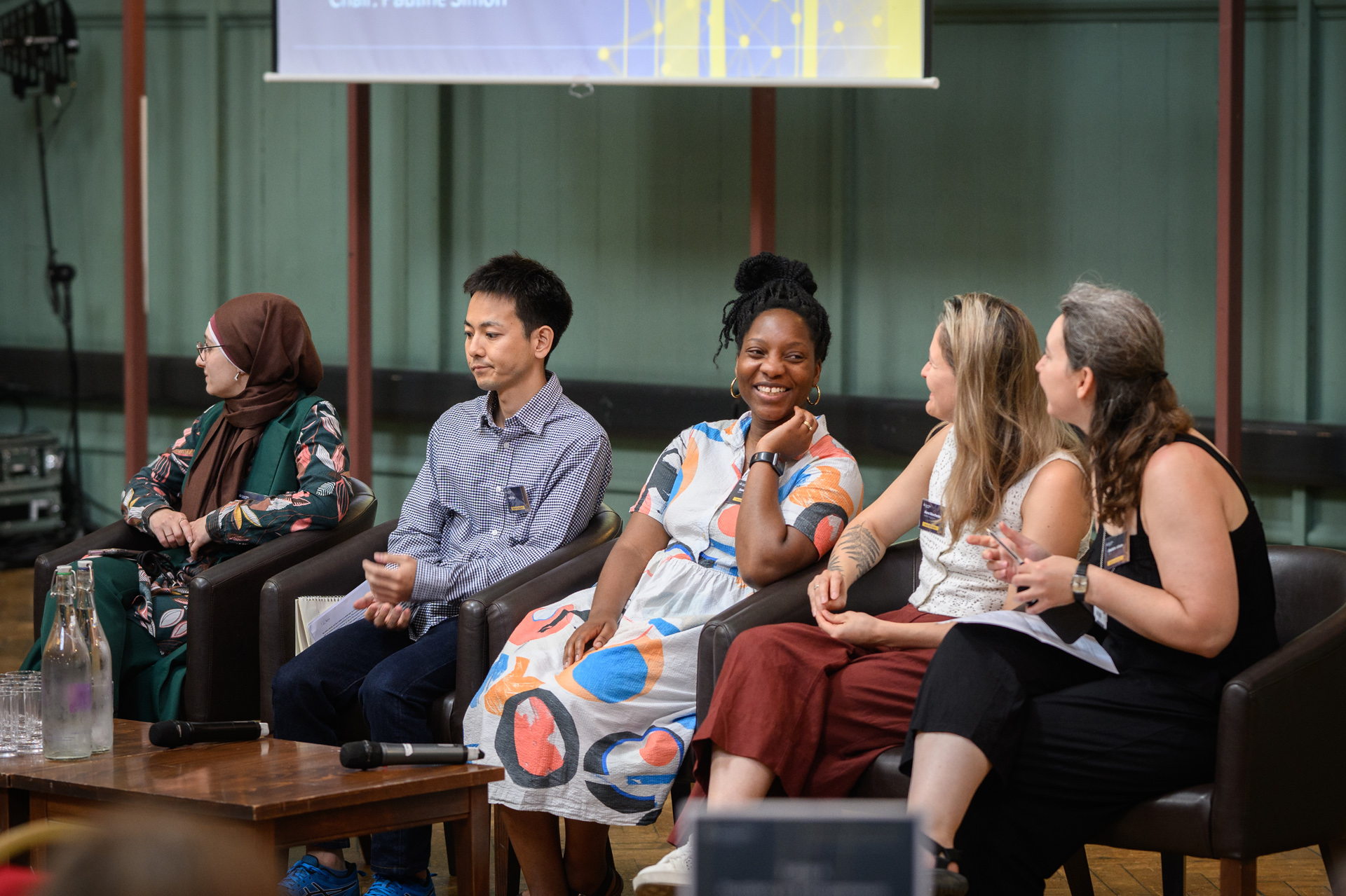 Five people sat in chairs, waiting to participate in a panel discussion at a conference. A black woman in the middle of the group is smiling to the women next to her.
