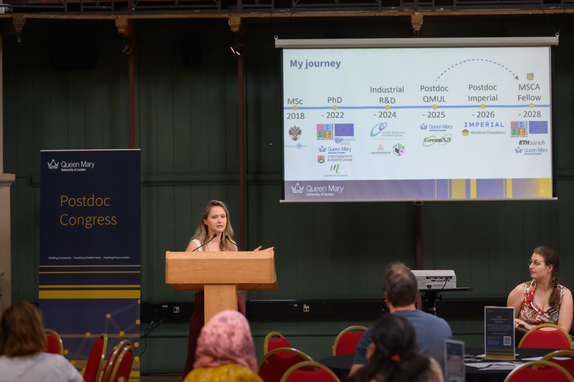 A woman standing at a podium, presenting at a conference, with presentation slides projected on to a screen to her right