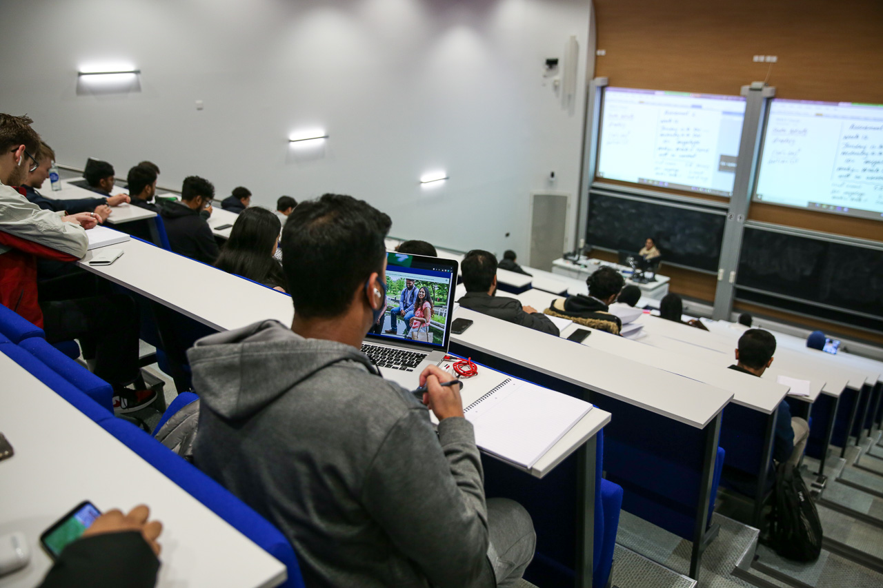 A student sitting at the back of a large tiered lecture theatre with a laptop on the table. The lecturer is very far away.